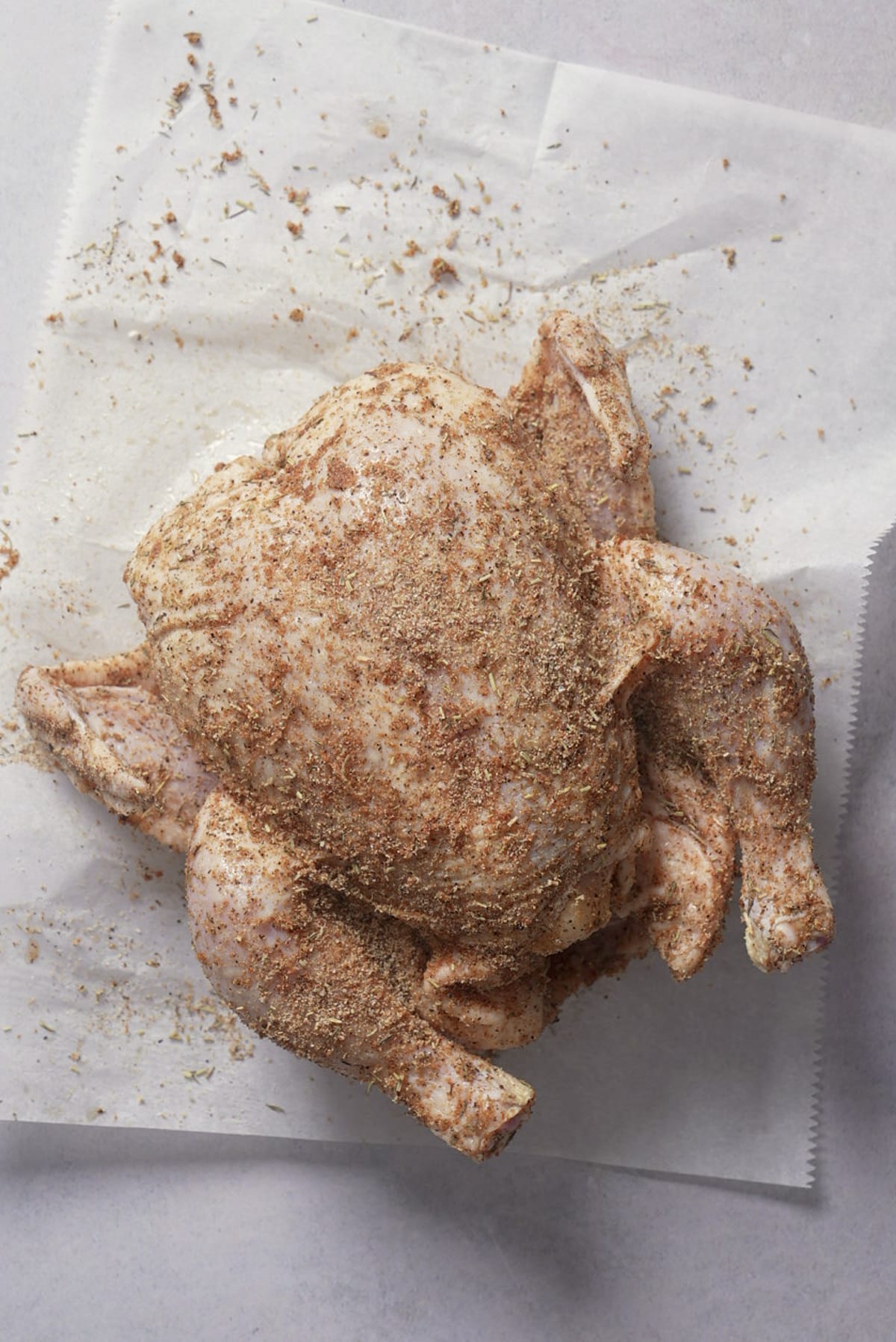 Whole raw chicken coated in a brown herb and spice rub, resting on parchment paper.