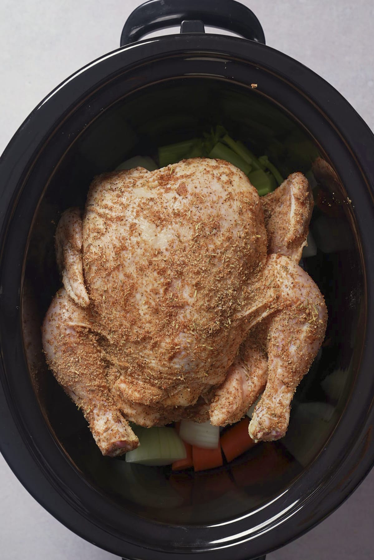 Seasoned raw whole chicken placed breast side up on a bed of onion, celery, and carrots inside a slow cooker.