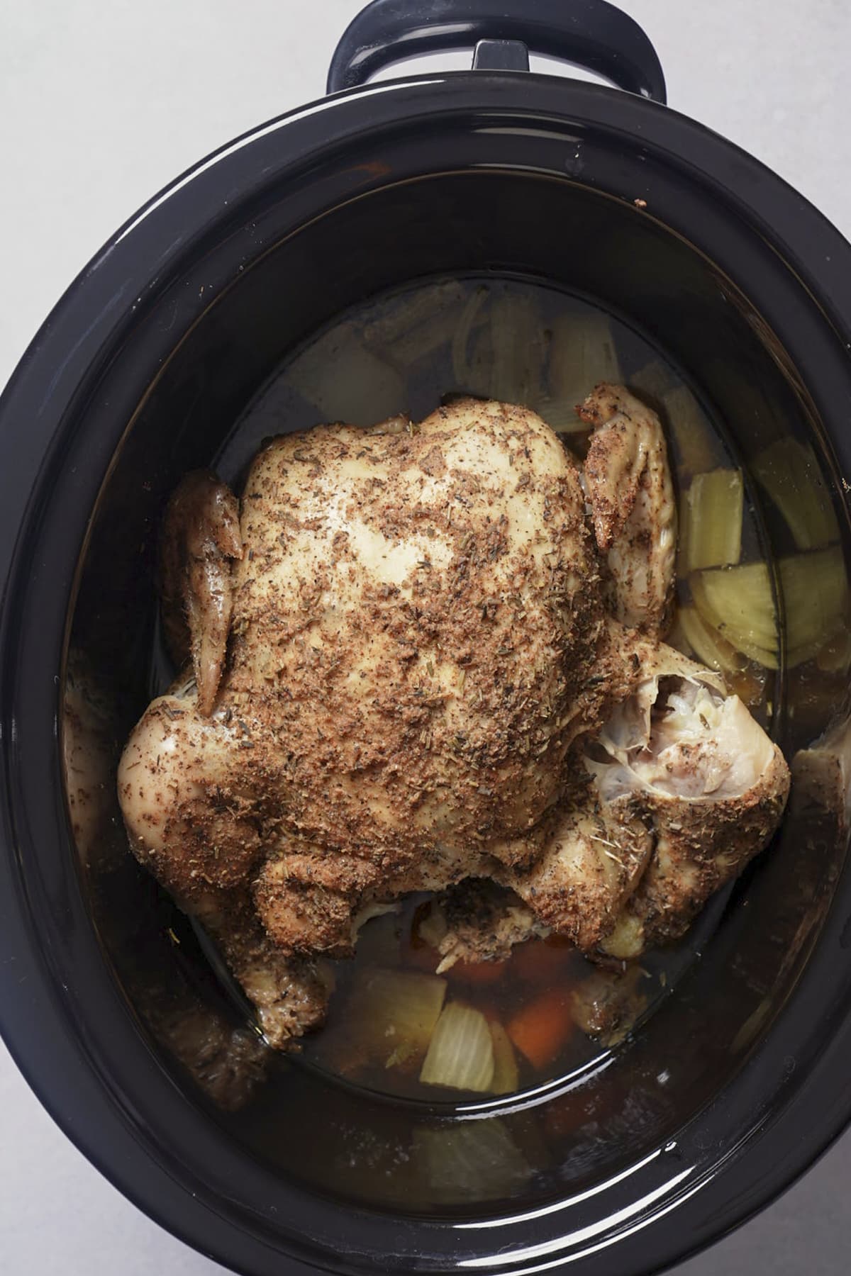 Fully cooked whole chicken in a slow cooker with browned seasoning and vegetables and juices around it.