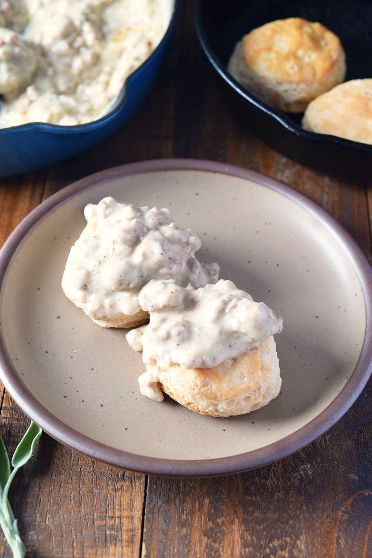Two biscuits topped with sausage gravy on a speckled plate, with the skillet of gravy and a pan of biscuits behind it.