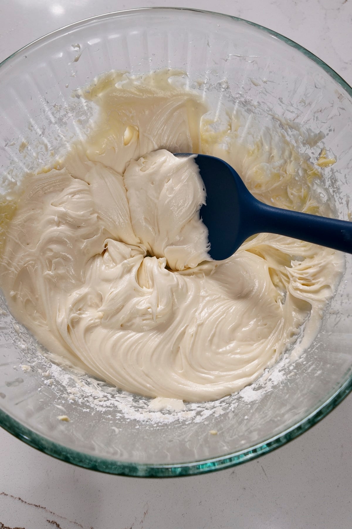cream cheese frosting in a glass bowl with a blue spatula sticking out.