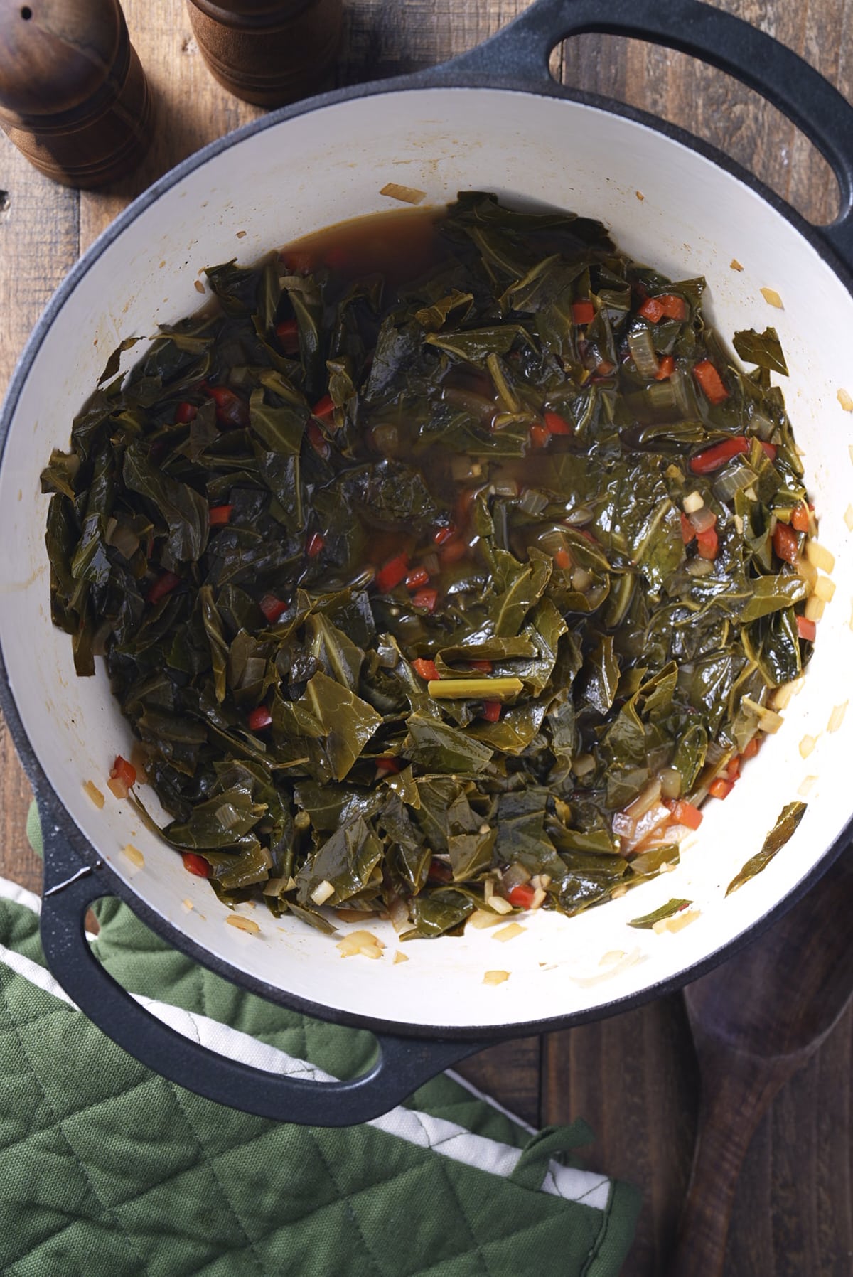 Finished collard greens and their pot likker in a white enameled Dutch oven on a wooden table, with a green cloth and wooden pepper mills alongside.
