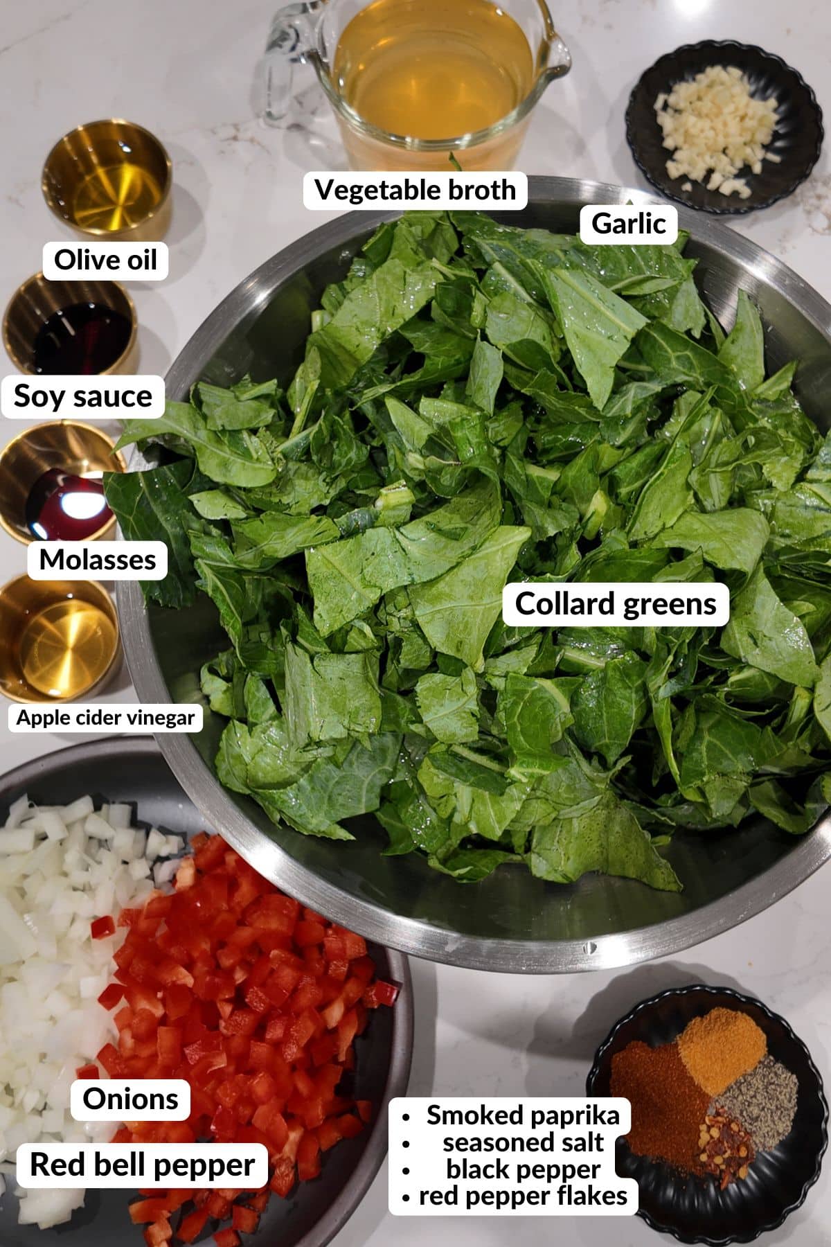 Labeled ingredients for meatless collard greens: a large bowl of chopped collard greens, a measuring cup of vegetable broth, minced garlic, olive oil, soy sauce, molasses, apple cider vinegar, diced onion and red bell pepper, and a dish of smoked paprika, seasoned salt, black pepper and red pepper flakes.