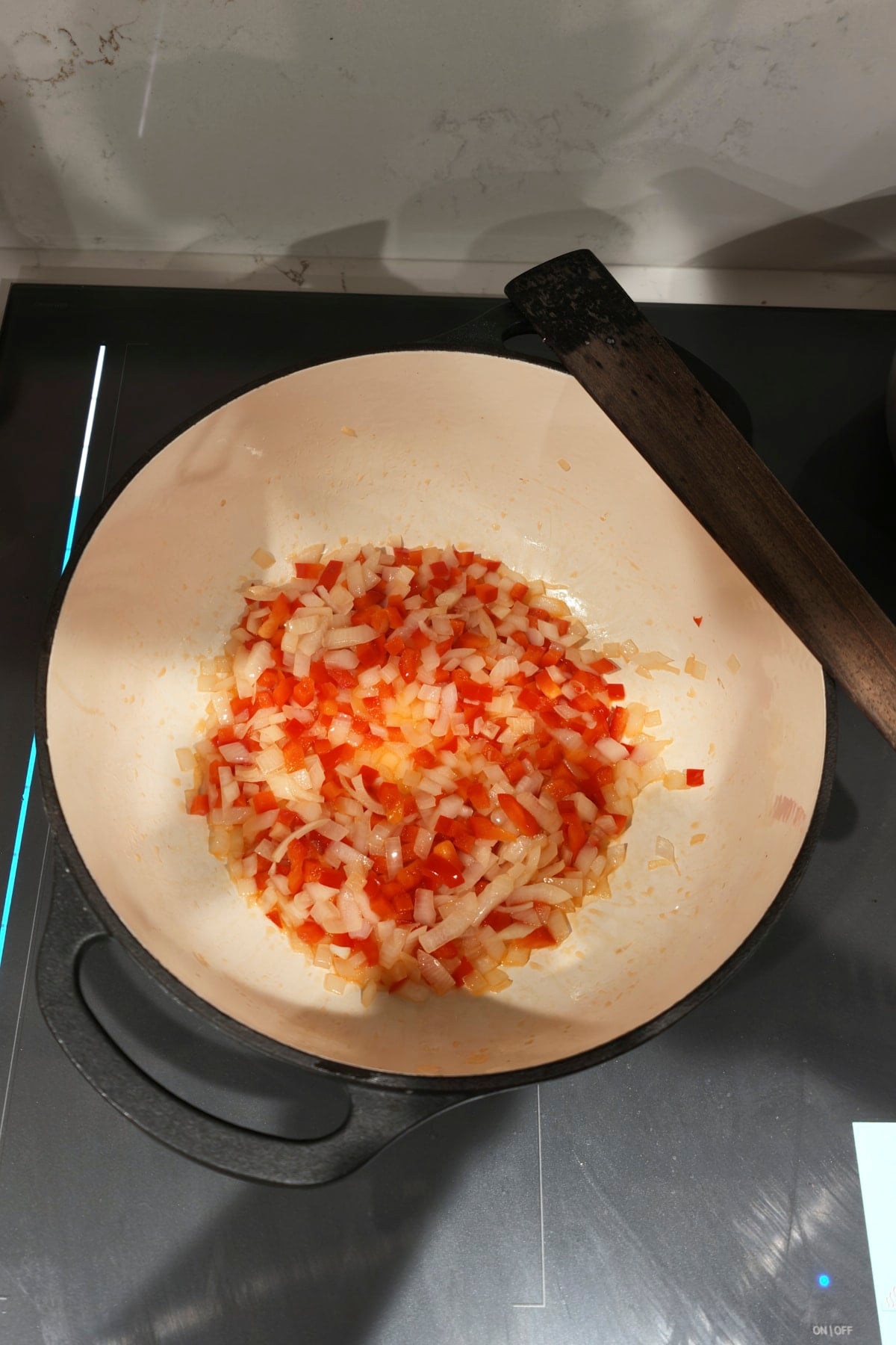 Diced white onion and red bell pepper softening in olive oil in a white enameled pot, with a wooden spoon resting on the rim.