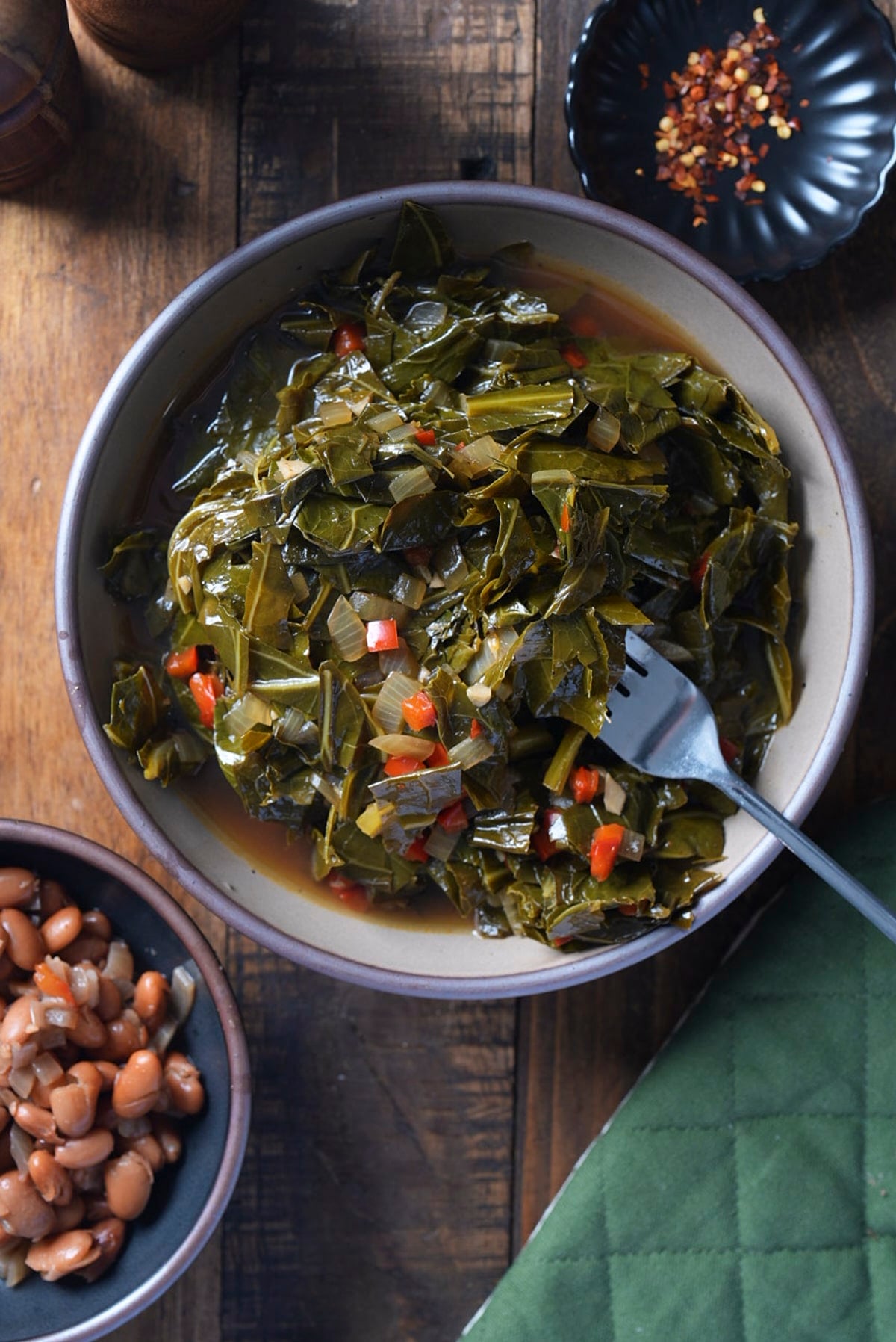 A bowl of collard greens sitting in pot likker with a fork resting in it, a bowl of pinto beans and a small dish of red pepper flakes alongside on a wooden table.