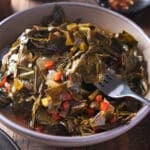 A heaped bowl of collard greens with a serving fork lifting some of the leaves, with a plate of white rice and a dish of red pepper flakes nearby.
