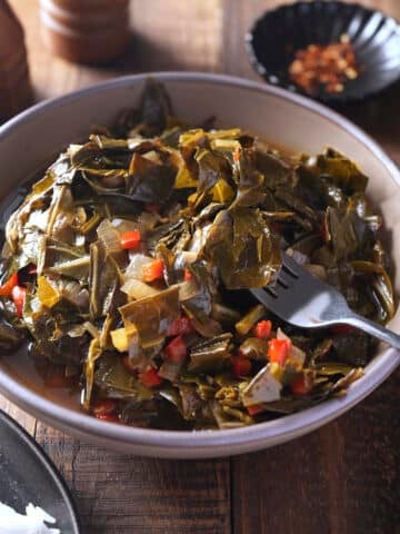 A heaped bowl of collard greens with a serving fork lifting some of the leaves, with a plate of white rice and a dish of red pepper flakes nearby.