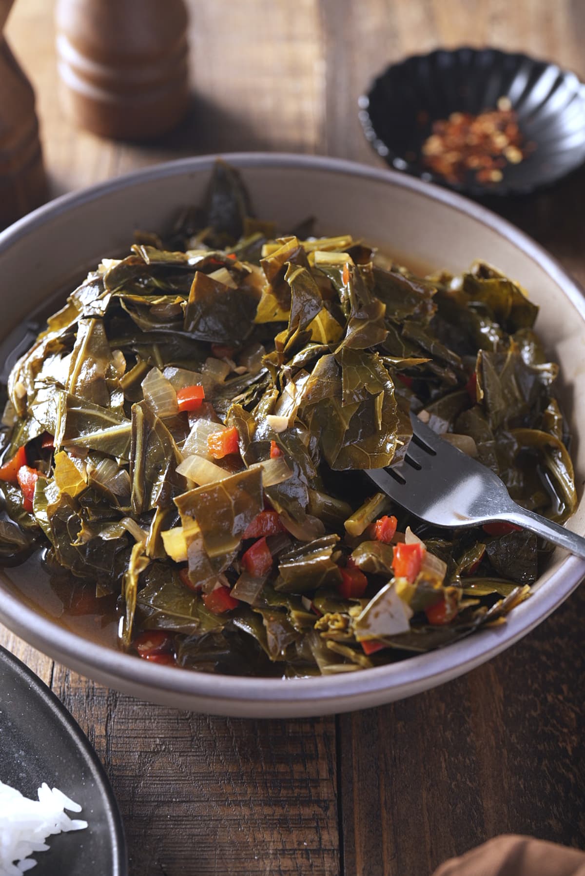 A heaped bowl of collard greens with a serving fork lifting some of the leaves, with a plate of white rice and a dish of red pepper flakes nearby.