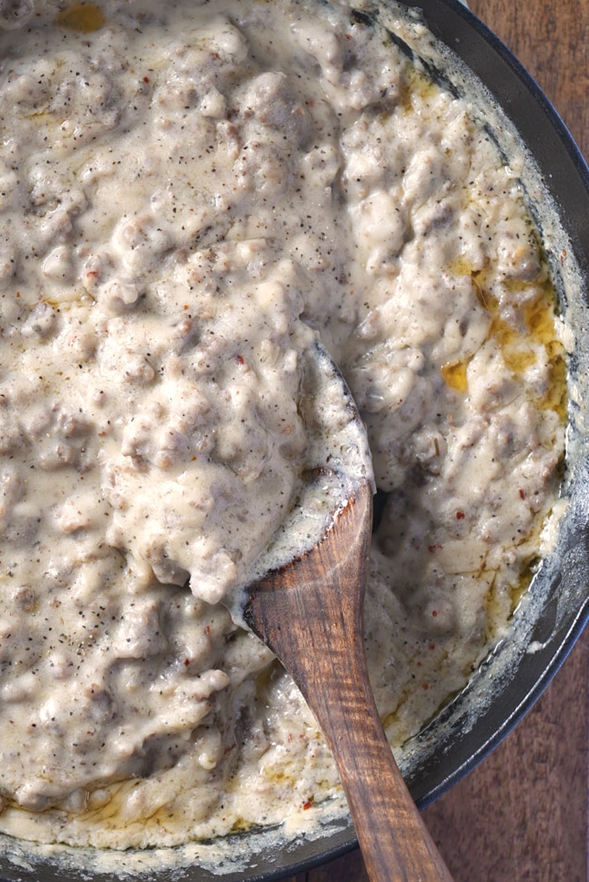 Close-up of chunky sausage gravy speckled with black pepper, scooped up on a wooden spoon in the skillet.