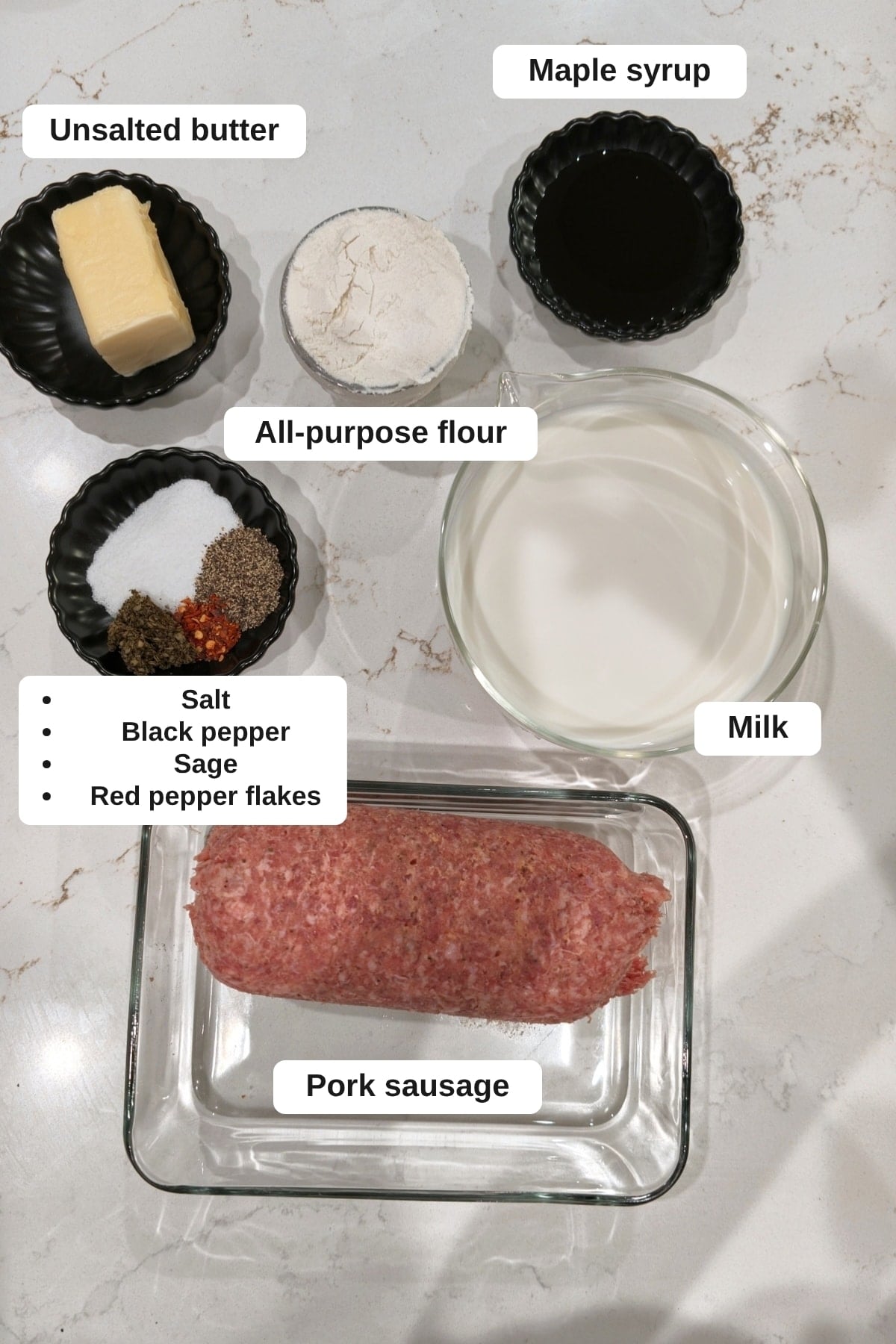Labeled sausage gravy ingredients on a marble counter: unsalted butter, all-purpose flour, maple syrup, milk, pork sausage, and a small bowl of salt, black pepper, sage and red pepper flakes.