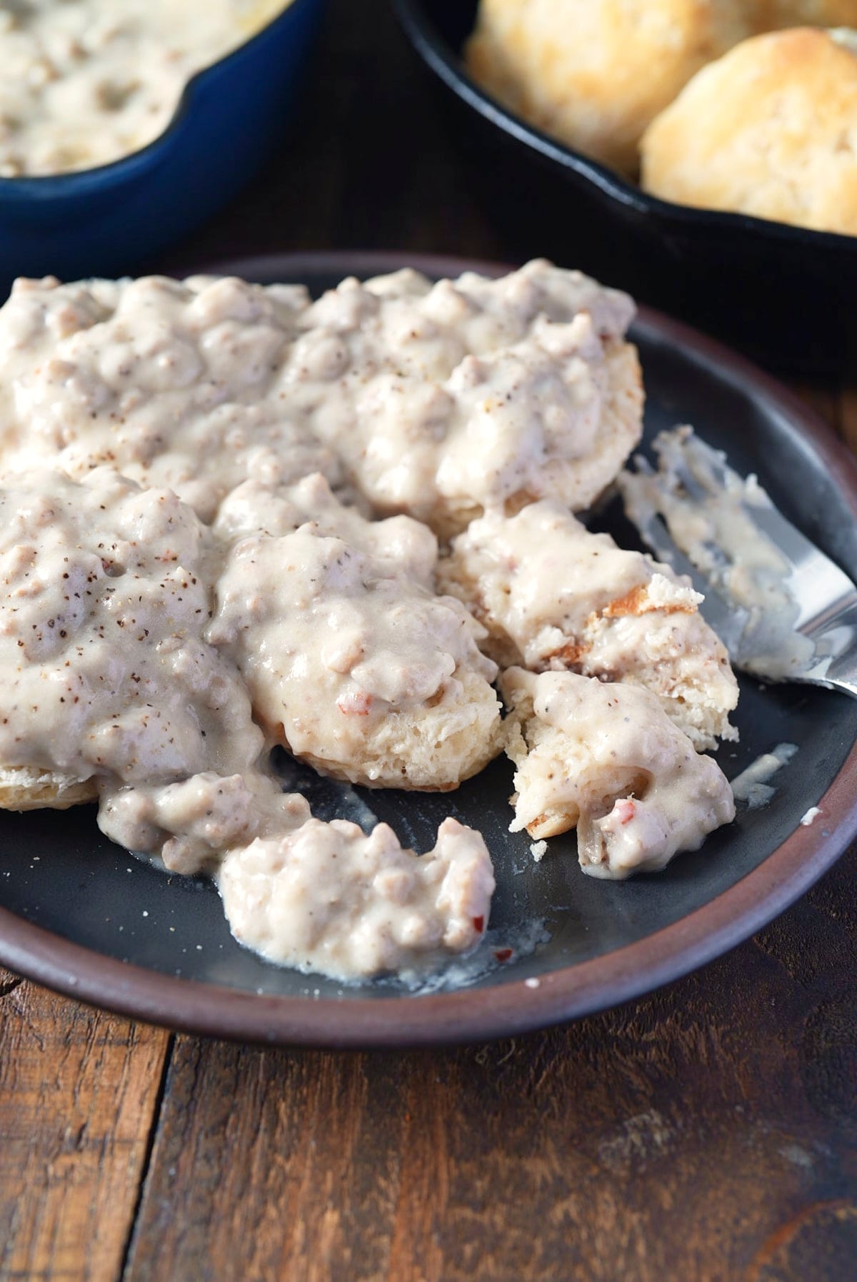 Split biscuits smothered in sausage gravy on a dark plate, with more biscuits and the skillet of gravy in the background.