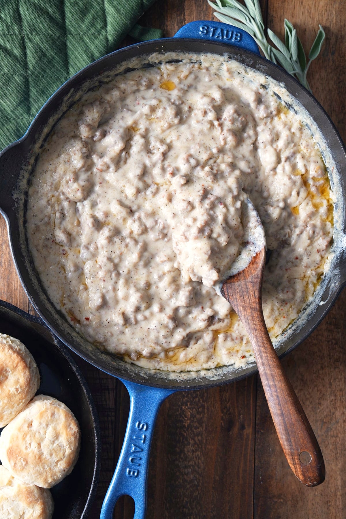 Overhead view of thick sausage gravy in a blue cast iron skillet with a wooden spoon, fresh sage sprigs on top, and biscuits on the side.