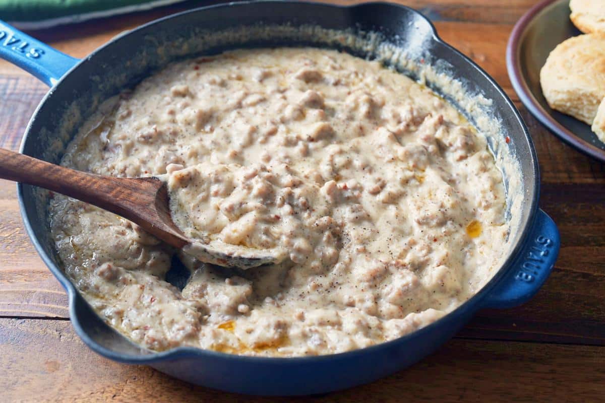 Side view of a blue cast iron skillet full of sausage gravy with a wooden spoon, and a plate of biscuits behind it.