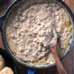 Creamy sausage gravy in a blue cast iron skillet with a wooden spoon resting in it, next to biscuits and fresh sage on a wooden table.