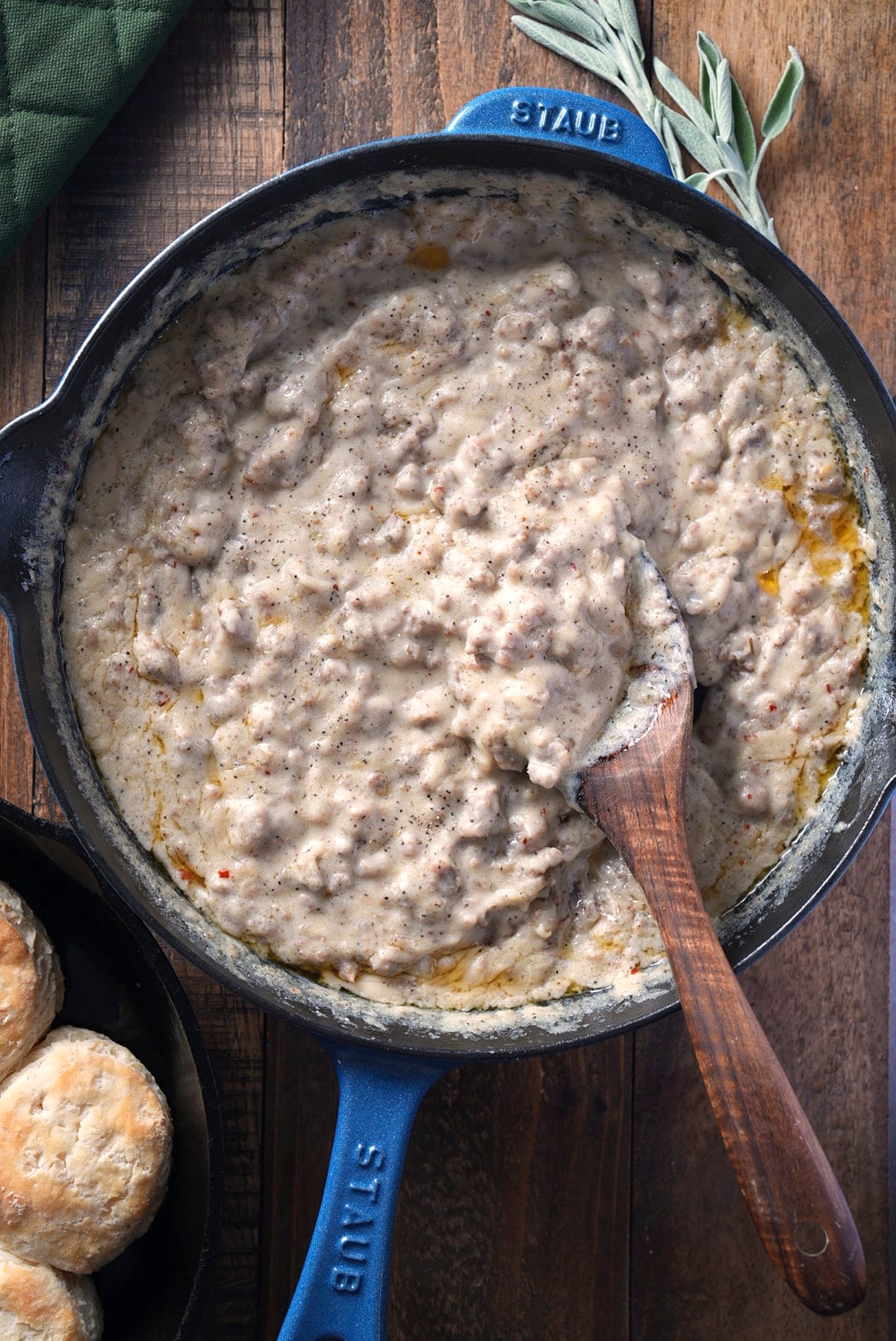 Creamy sausage gravy in a blue cast iron skillet with a wooden spoon resting in it, next to biscuits and fresh sage on a wooden table.