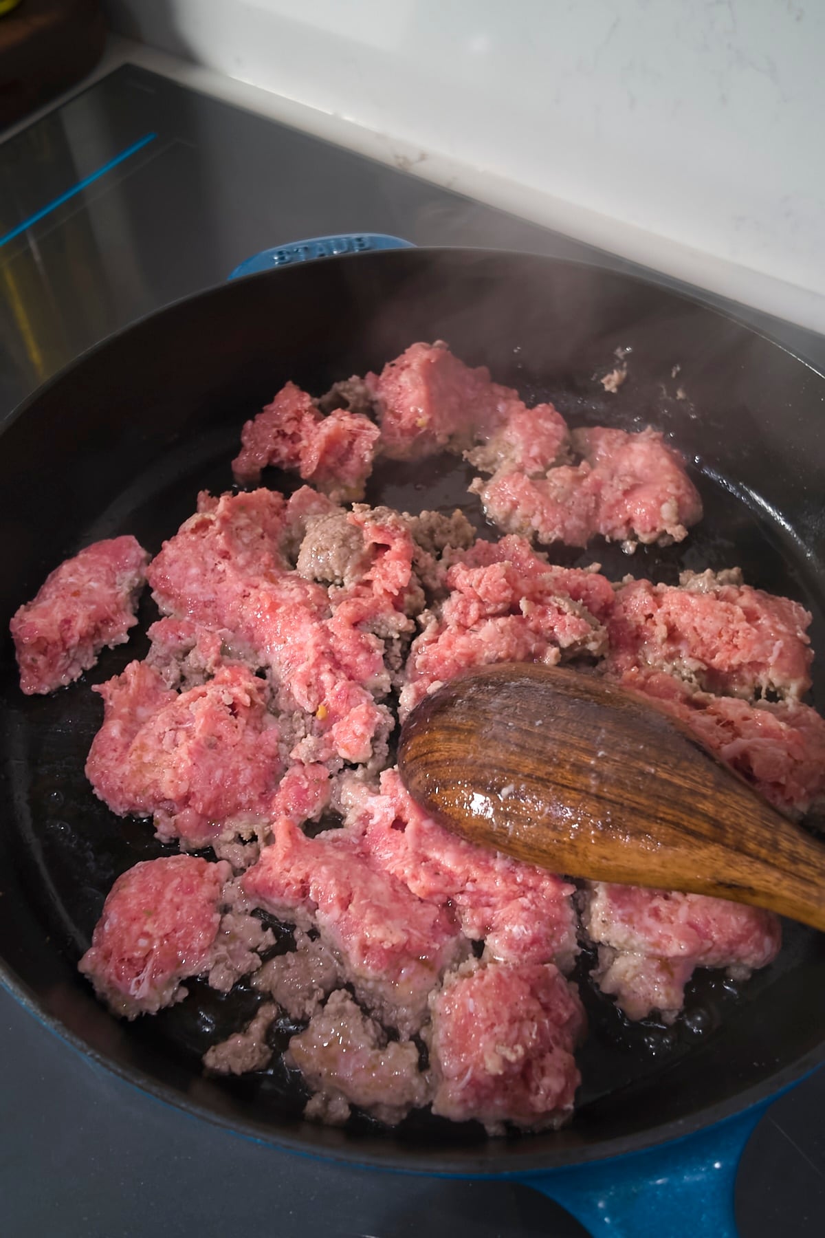 Raw pork sausage being broken apart with a wooden spoon in a blue cast iron skillet.