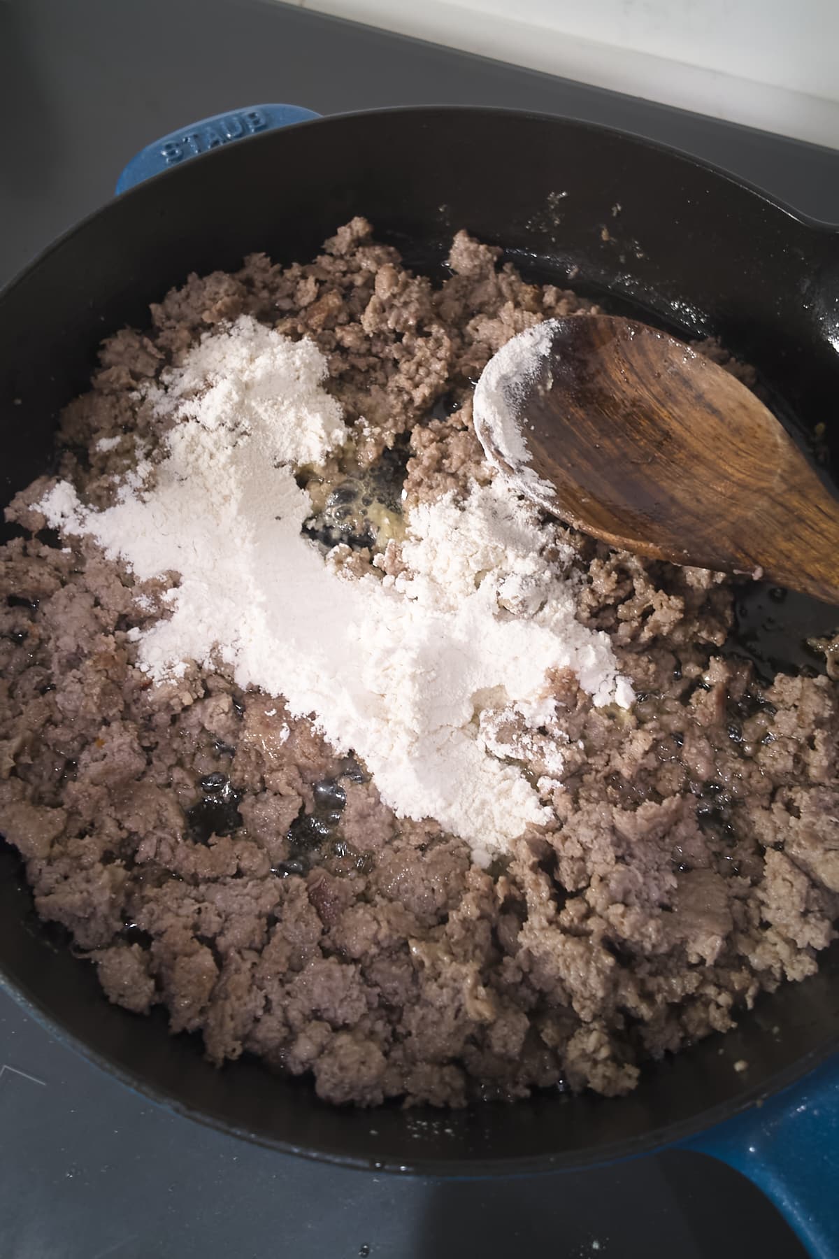 Flour sprinkled over browned, crumbled sausage in a cast iron skillet, with a wooden spoon ready to stir.