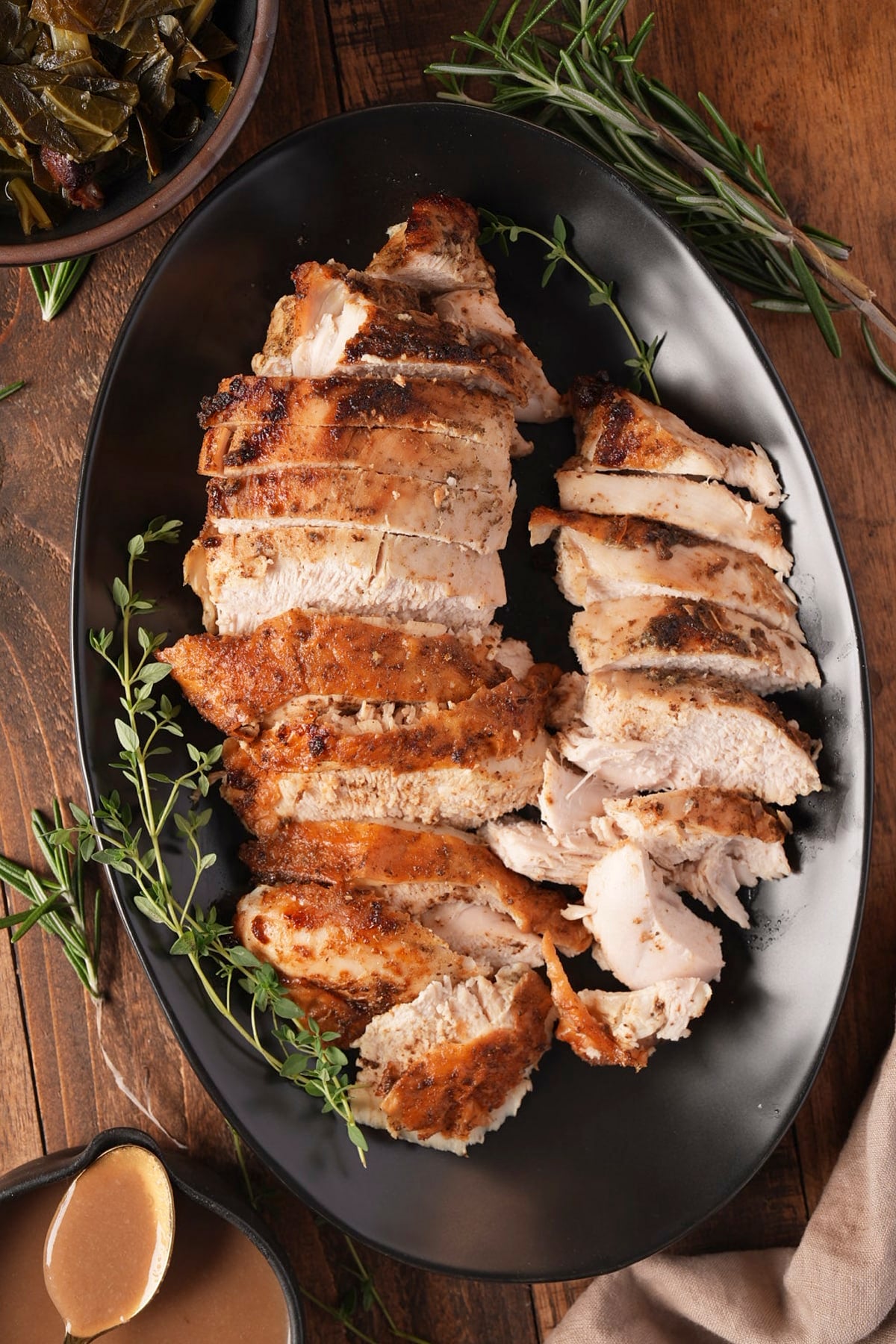 Sliced turkey breast with golden, seasoned skin on a black platter, with gravy and collard greens on the side
