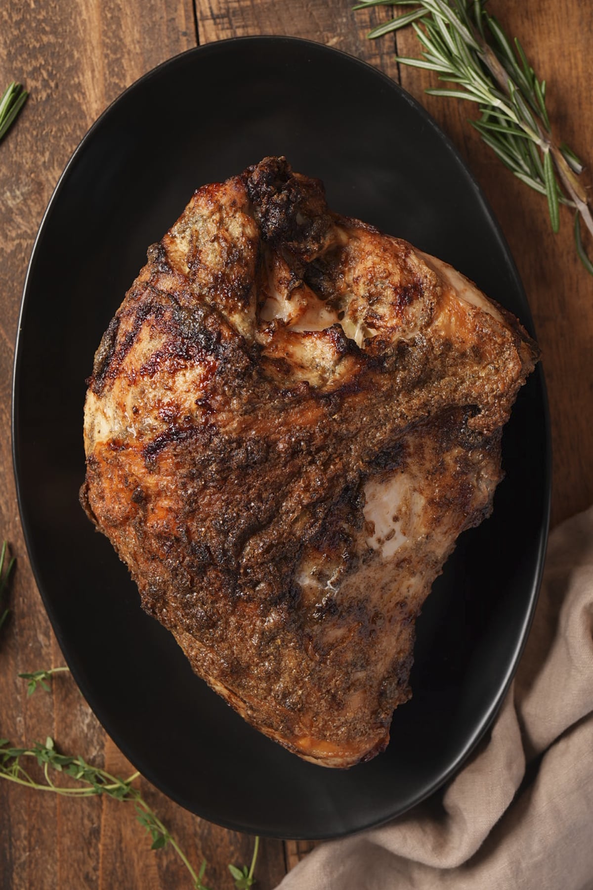Whole golden brown turkey breast on a black platter with fresh rosemary and thyme