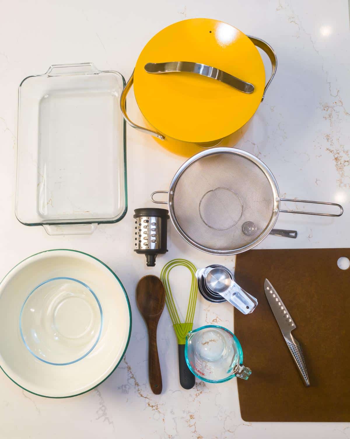 Equipment for the recipe laid out on a counter, including a glass baking dish, a pot, a colander, a grater, a whisk, and measuring cups.