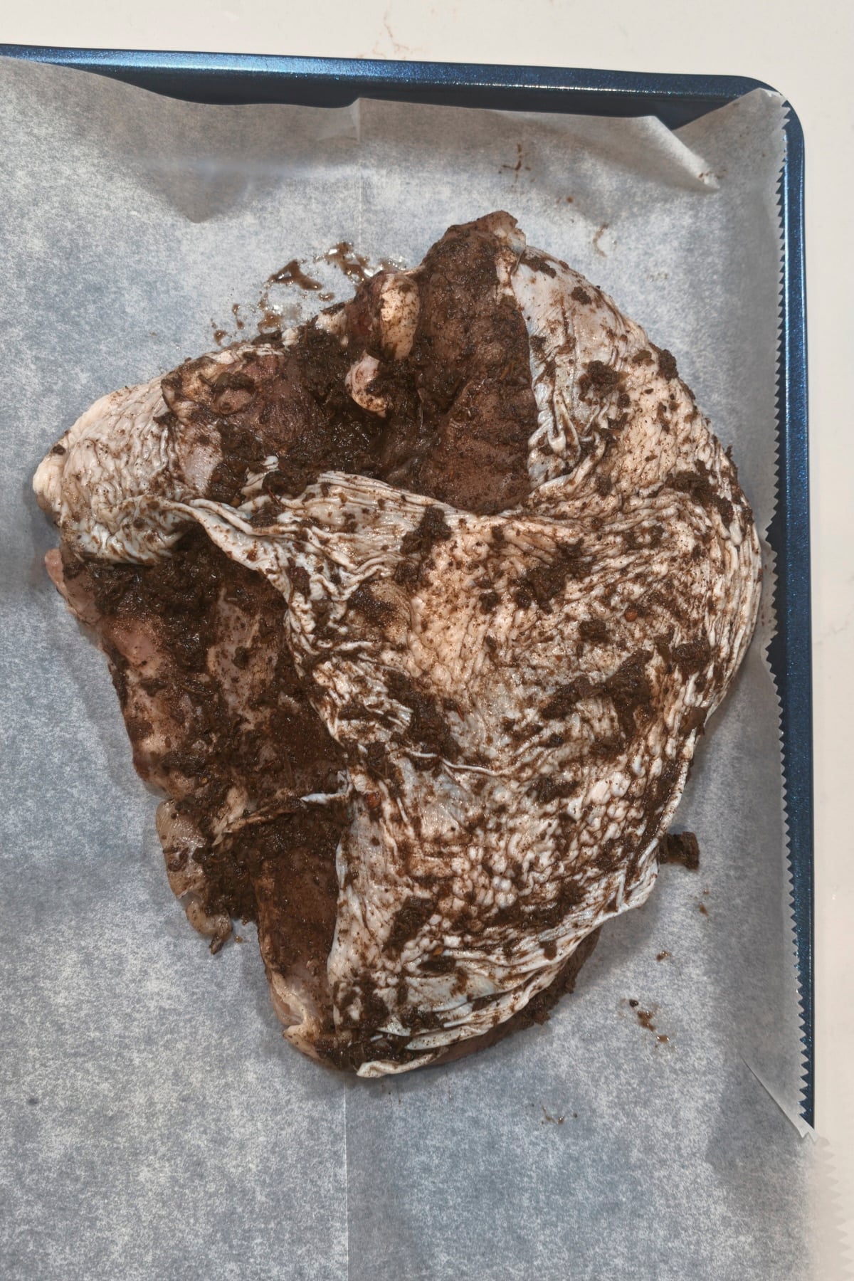 Jerk paste being spread over the skin of a raw turkey breast on a parchment-lined pan.