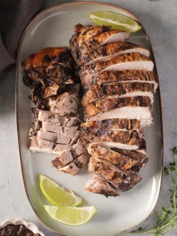 Jerk turkey breast carved into slices on a white platter with lime wedges, showing white meat under a dark seasoned crust.
