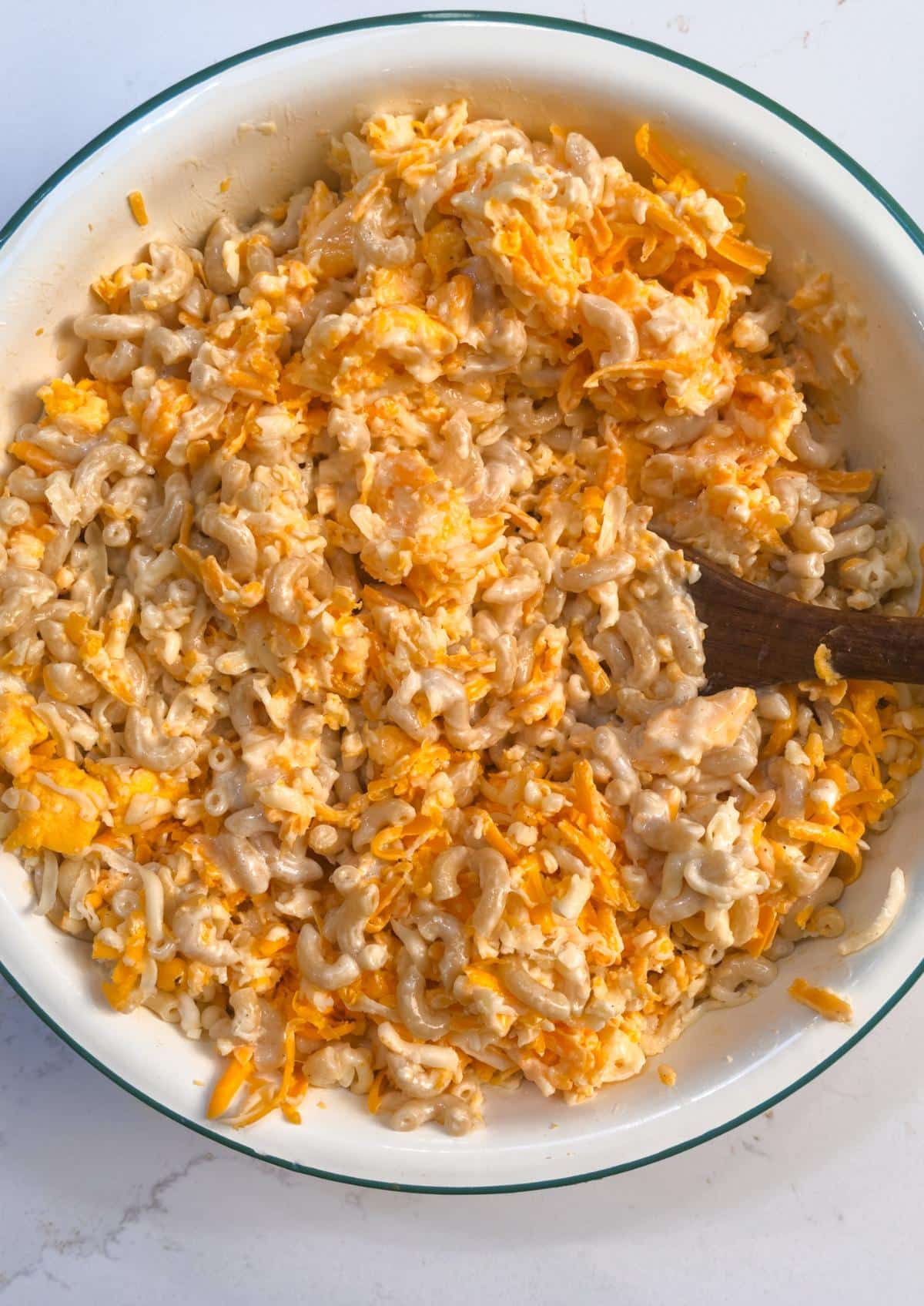 Shredded cheese stirred into the macaroni in a large white bowl with a wooden spoon.