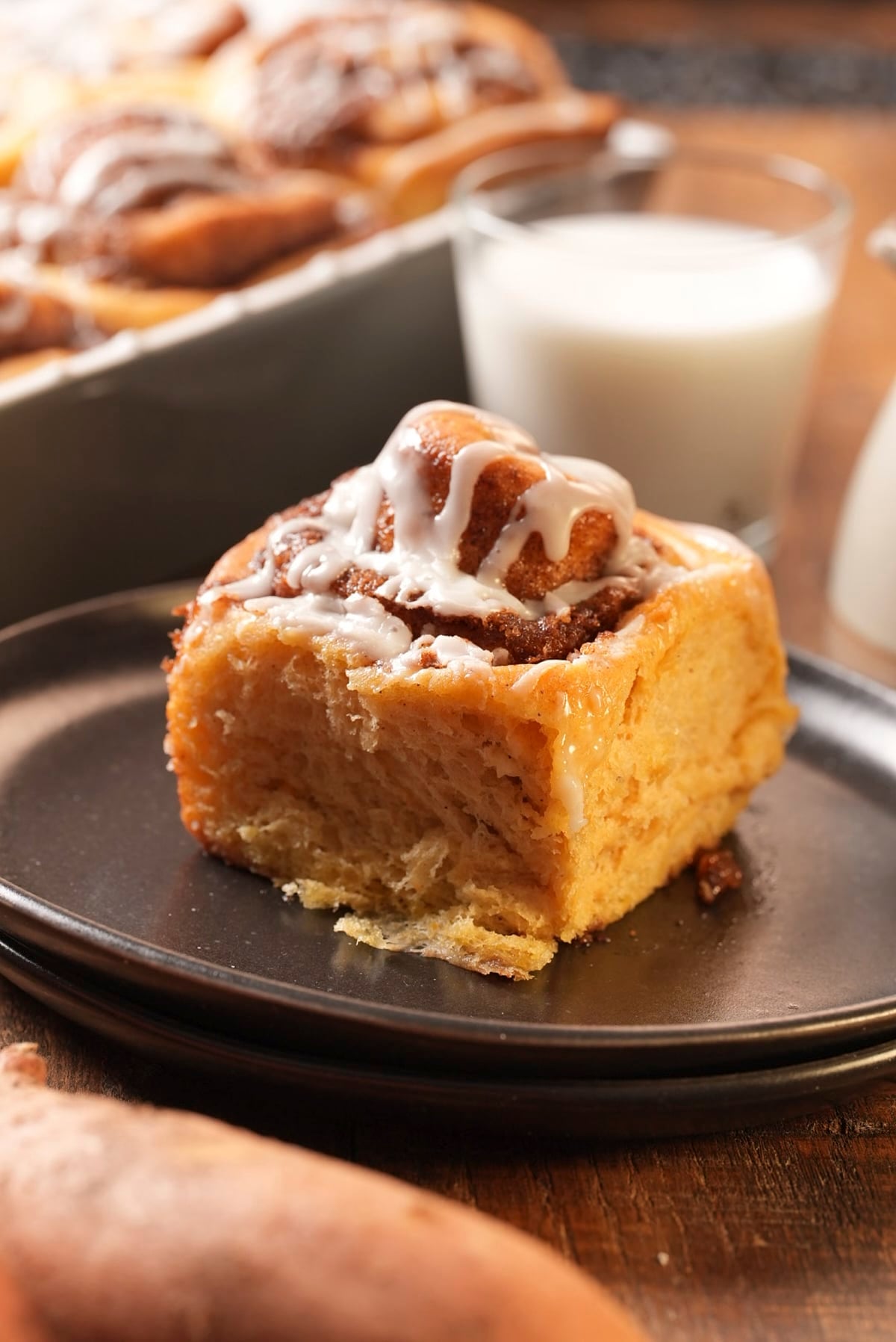 One glazed sweet potato cinnamon roll on a dark plate with a glass of milk and the pan behind it.